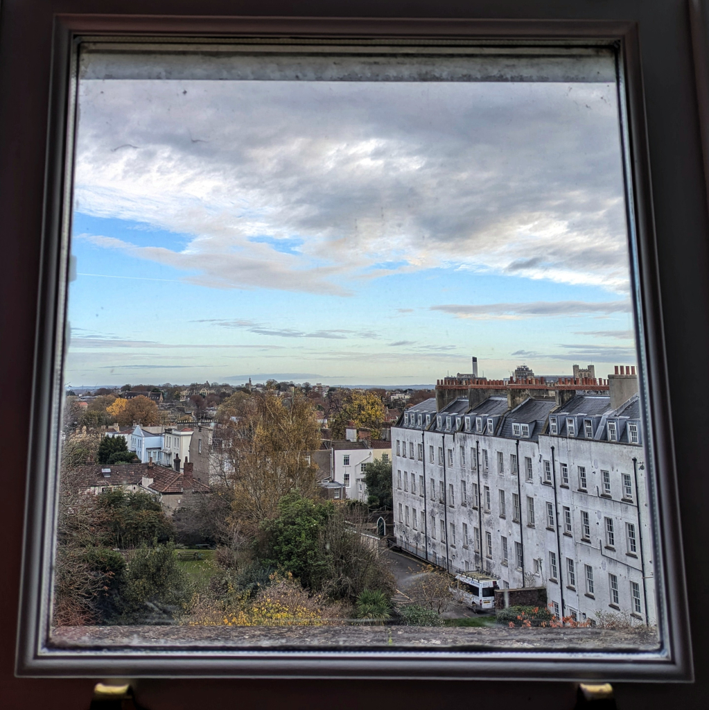 Looking through a window in the early morning over a large garden and houses. The light is soft and gray. Shredded clouds are moving slowly across a light blue sky. 