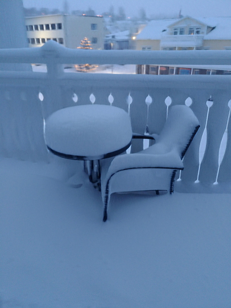 Image of a wooden balcony with round black metal table and chair. Top rail of balcony, table and chairs are covered with a thick layer of snow. Table has a pristine layer snow on it about 30cm deep which looks like an enormous white cake. In the distance are cream coloured buildings and a tree with white Christmas lights. Picture was taken in Akureyri in Northern Iceland.