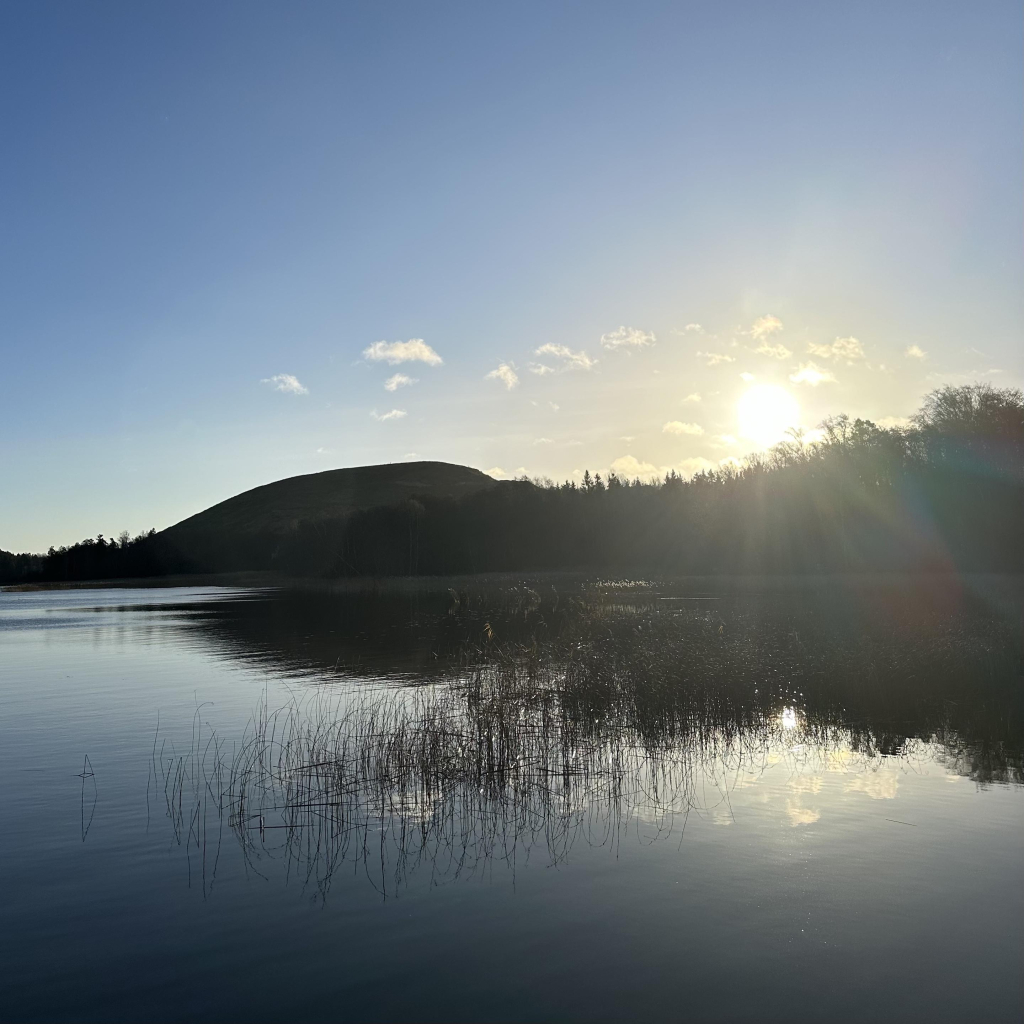 View over a lake with small puffy white clouds and a sun shining bright making the forest and a hill on the other side into dark silhouettes.