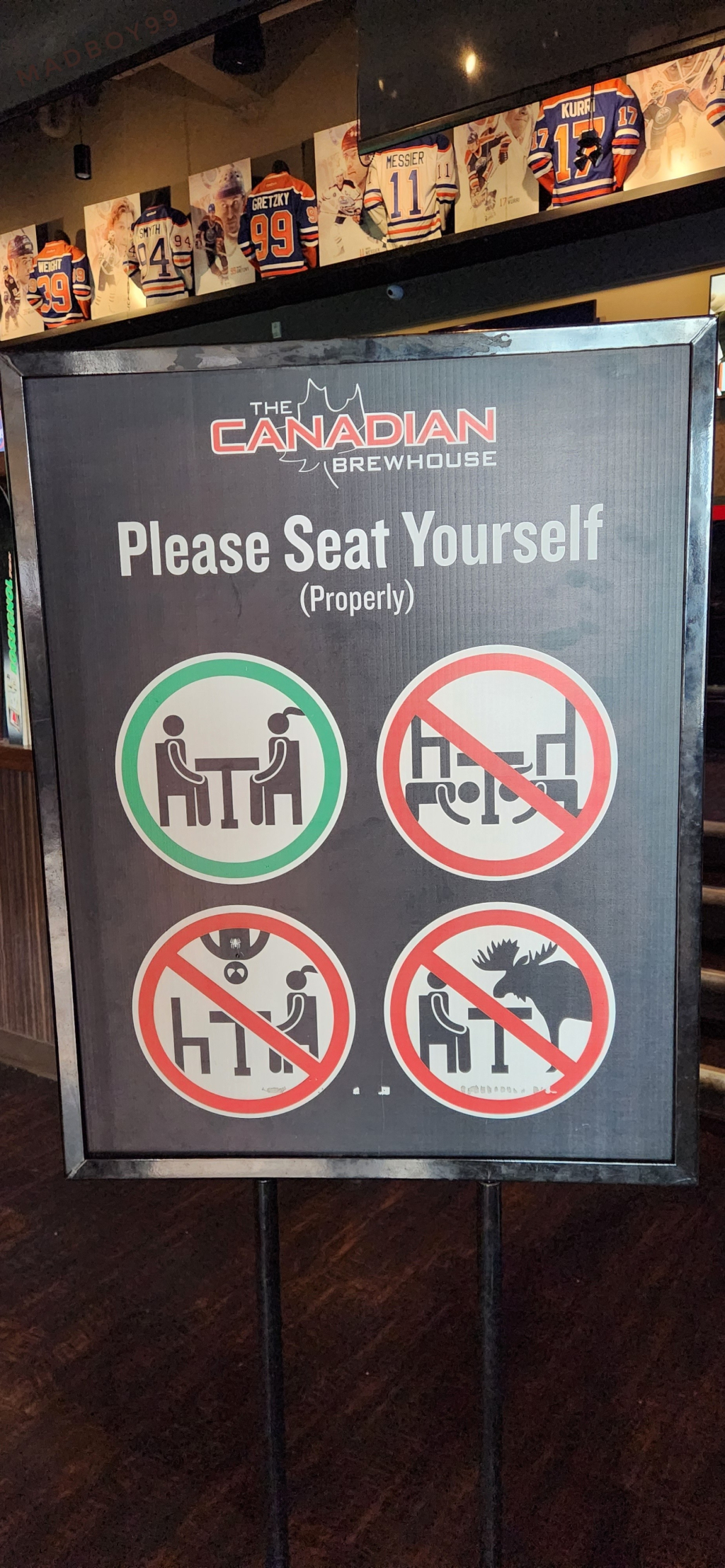 A seating sign at a restaurant entrance that reads Please seat yourself (properly), 4 drawings one has 2 people sitting properly, other 3 not so, one under the table, another on the ceiling, last one with a moose.