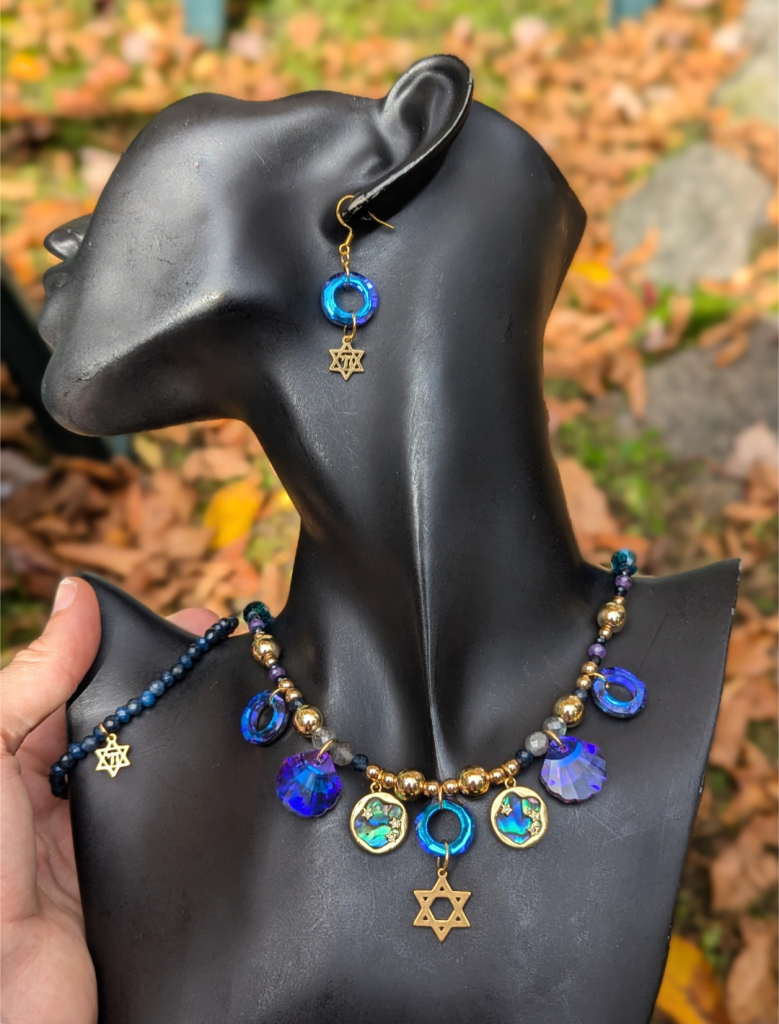 On a beautiful black jewelry show doll, a beaded blue gold set with a handmade necklace of Magen David pendant, faceted glass round and she'll shaped purple blue pendants, golden abalone shell pendant, beaded sapphire bracelet with Chai Magen David bracelet and fitting loop Magen David earrings 
Vermont fall leaves in the background