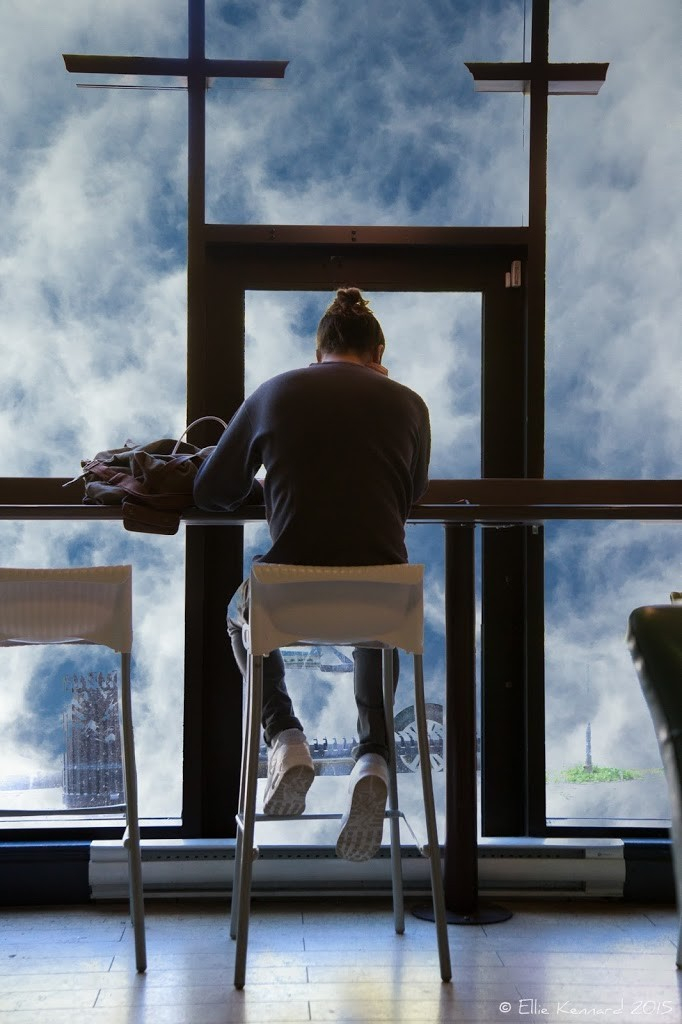 Head in the clouds.  A man with a bun is sitting on a high stool with his back to us, facing a window. He is in a cafe but appears to be in the clouds from the scene outside the window.