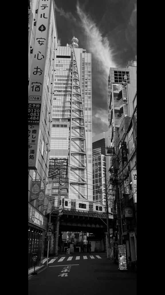 A 65x24 vertical aspect ratio photo made with the 65x24 app on an iPhone 12Pro. 

We are in Shimbashi, a gritty neighbourhood of Tokyo full of bars, restaurants and a fave for workers to visit once their day is done. 

I was in one of the narrow streets of restaurants and bars, looking towards the skyscrapers of the neighbouring Shiodome district. One large, distinctive tall building is in the middle of the background. Large metal struts dominate the outside of the building. This is the Nittele Tower building (Nippon Television Tower). 32 storeys high, Nittele was designed by the Richard Rodgers Consortium. 

In the foreground the word "To-ma-re" (Stop in Japanese) is painted on the road. A train of the Yamanote Line crosses the bridge in the middle-ground of the photo. 