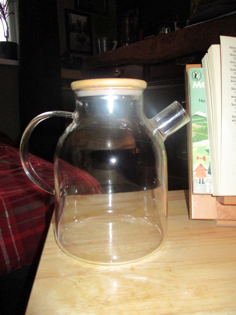 A glass teapot on a table next to a desktop lectern with Moomin books on it.  Various plants, a chest of drawers and other tat in the background.