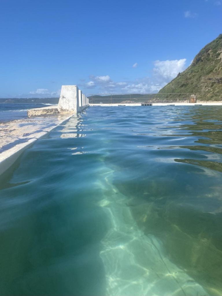 Taken while swimming on a sunny day and looking along the weathered white back wall of the Merewether Ocean Baths with the concrete lap lane markers visible from the side. The water is a very pale turquoise over the white painted edge of the pool floor with light refraction patterns. It's a darker blue green in the rest of the pool.