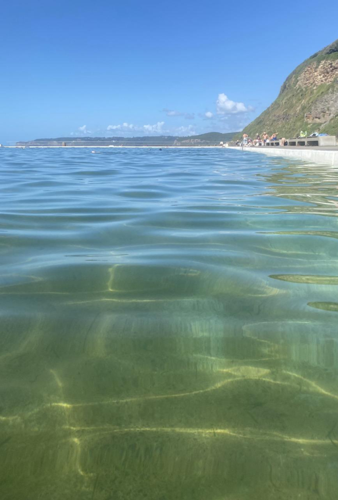 Taken while swimming and looking southwards across the light filled water to the distant curve of coastline with blue sky and little puffy clouds above those hills. In the foreground the water is light green with yellow highlights where the refracted light is making patterns.  