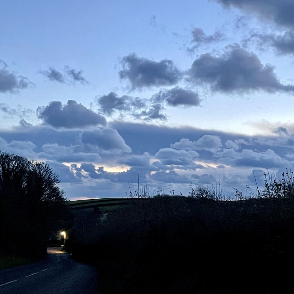 Dusky evening sky with cumulo nimbus clouds in slate blue tones with a hint of golden sunshine behind, over a foreground country road in darkness with silhouetted hills in the mid ground. 