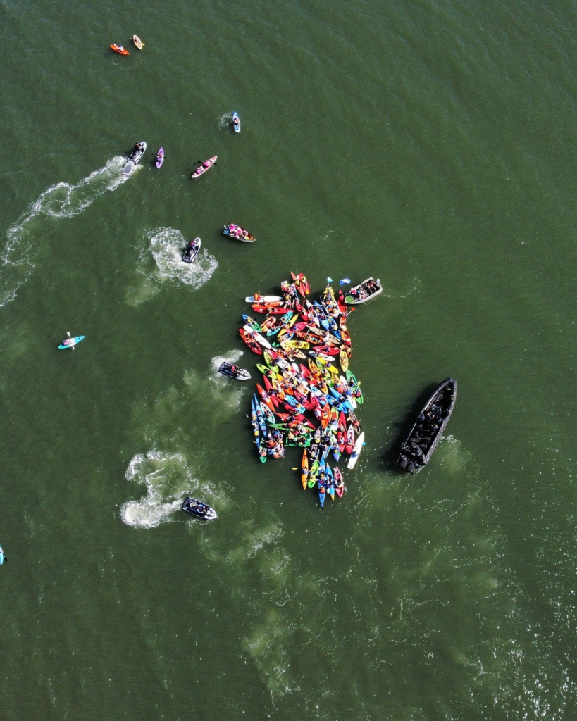 An image taken from Rising Tide's Facebook page. It shows 100 people in kayaks out on the water at Newcastle coal port (in New South Wales, Australia) being confronted with a police vessel filled with officers. The water is murky green, the kayaks are very brightly coloured (red, yellow, green, blue, etc) and the police boat is darkest black.