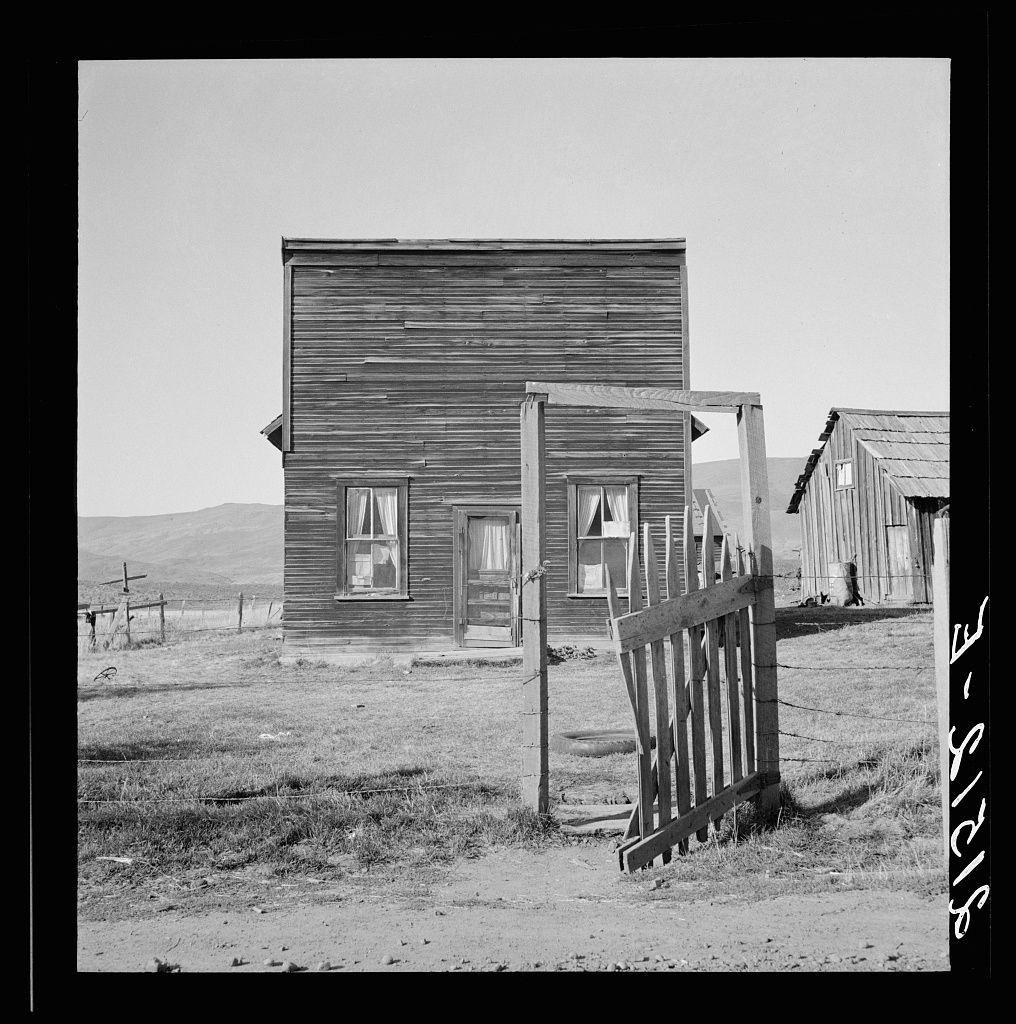 The image depicts a black and white photograph of an old, rustic wooden building with multiple windows. The structure appears to be situated in a rural area with barren ground surrounding it, indicating arid or semi-arid conditions typical of farmland or desert regions.

In the background, there are additional dilapidated structures that seem similar in construction style but less maintained than the main house, possibly suggesting an old settlement or farmstead. A broken wooden fence is visible in front of the building with a gate partially standing on its side, adding to the sense of neglect and abandonment.

The overall atmosphere conveyed by this photograph suggests desolation and decay typical for ghost towns during periods where economic activity ceased or drastically declined. The lack of vegetation and the arid landscape indicate harsh environmental conditions that may have contributed to the downfall of such settlements over time.