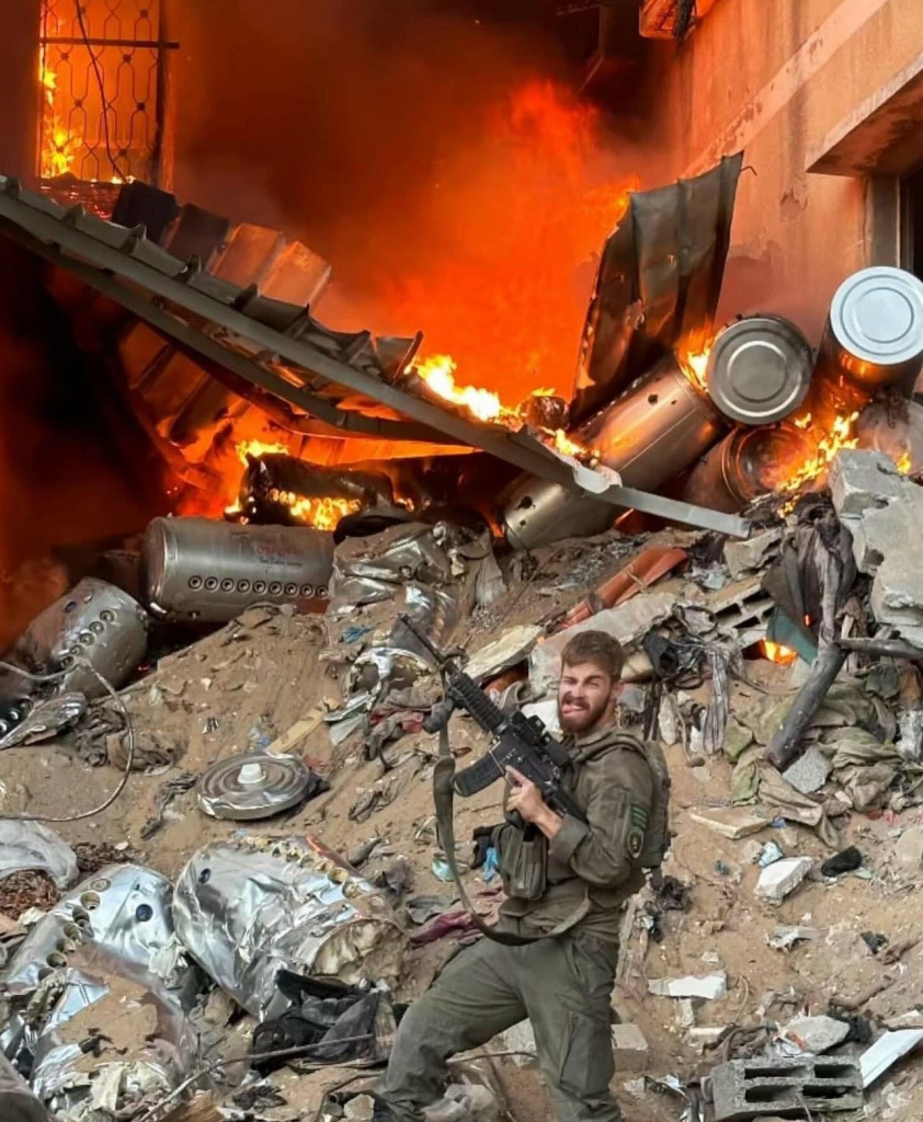 Ein israelischer Soldat vor einem Geröllhaufen aus zerstörten Trümmerteilen, Bauschutt und Dreck, im Hintergrund brennt ein Feuer. Der Soldat hält sein Waffe in die Höhe, streckt die Zunge raus und grinst in die Kamera. 