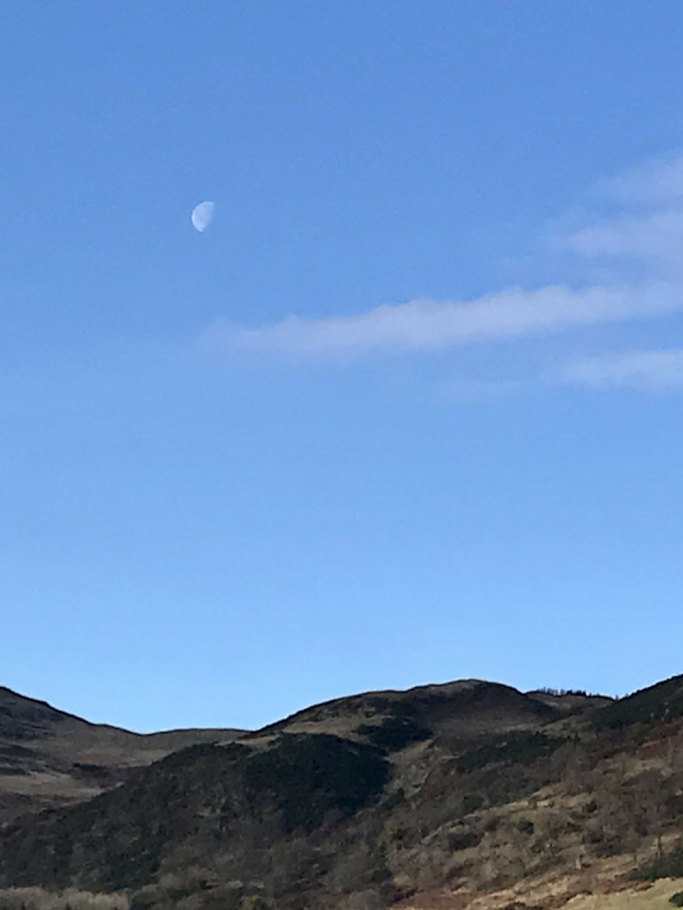 tops of rugged hills reach for a blue sky with just a few wispy watercolour clouds pushing into the frame from the right, seemingly indicating the presence of a half-moon.