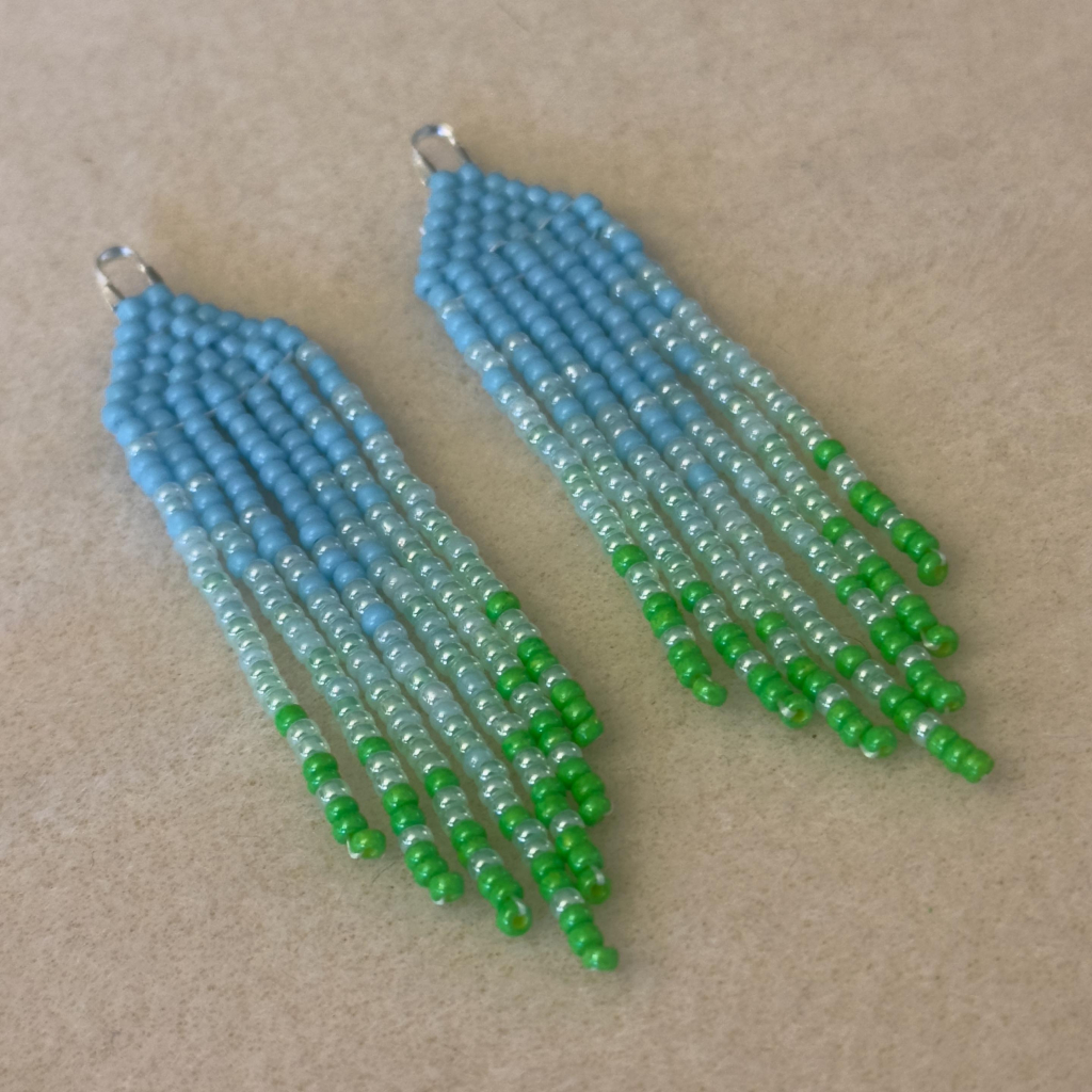 A pair of blue to green ombre mini beaded fringe earrings on a bead mat