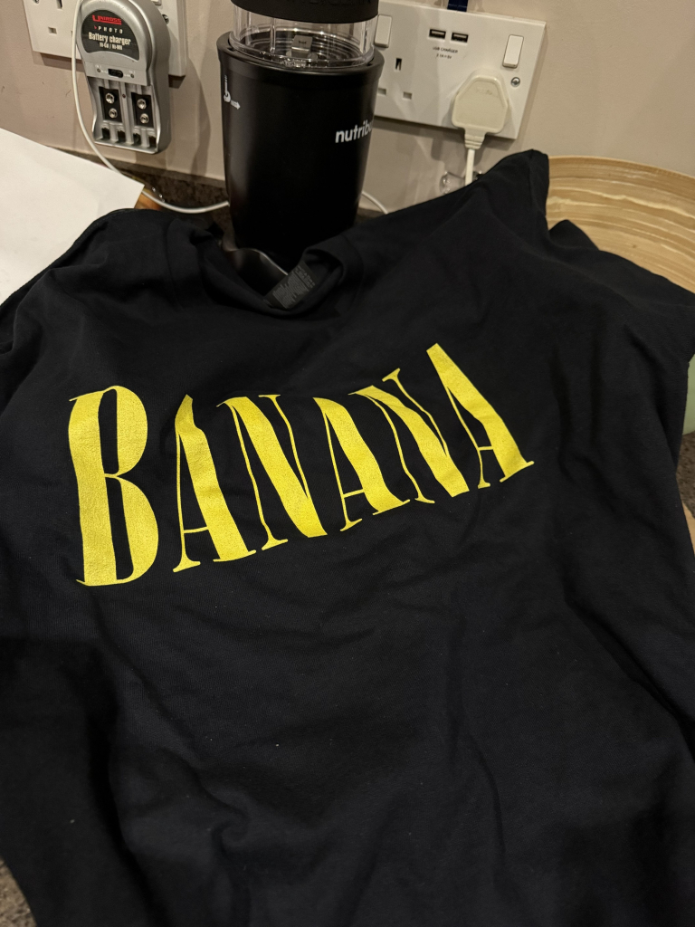 A black t-shirt with the word "BANANA" printed in bold yellow letters. In the background, a blender and a power outlet are visible.