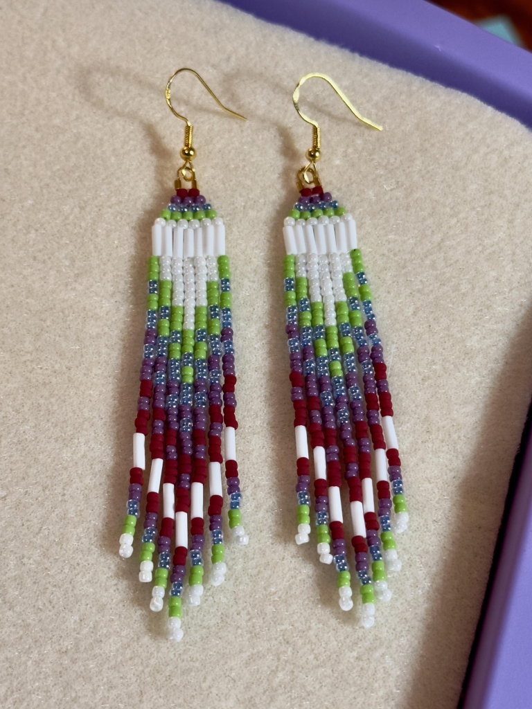 A pair of pearl white, apple green, blue, puprle, and matte dark red beaded fringe earrings with gold hooks on a cream colored bead mat.