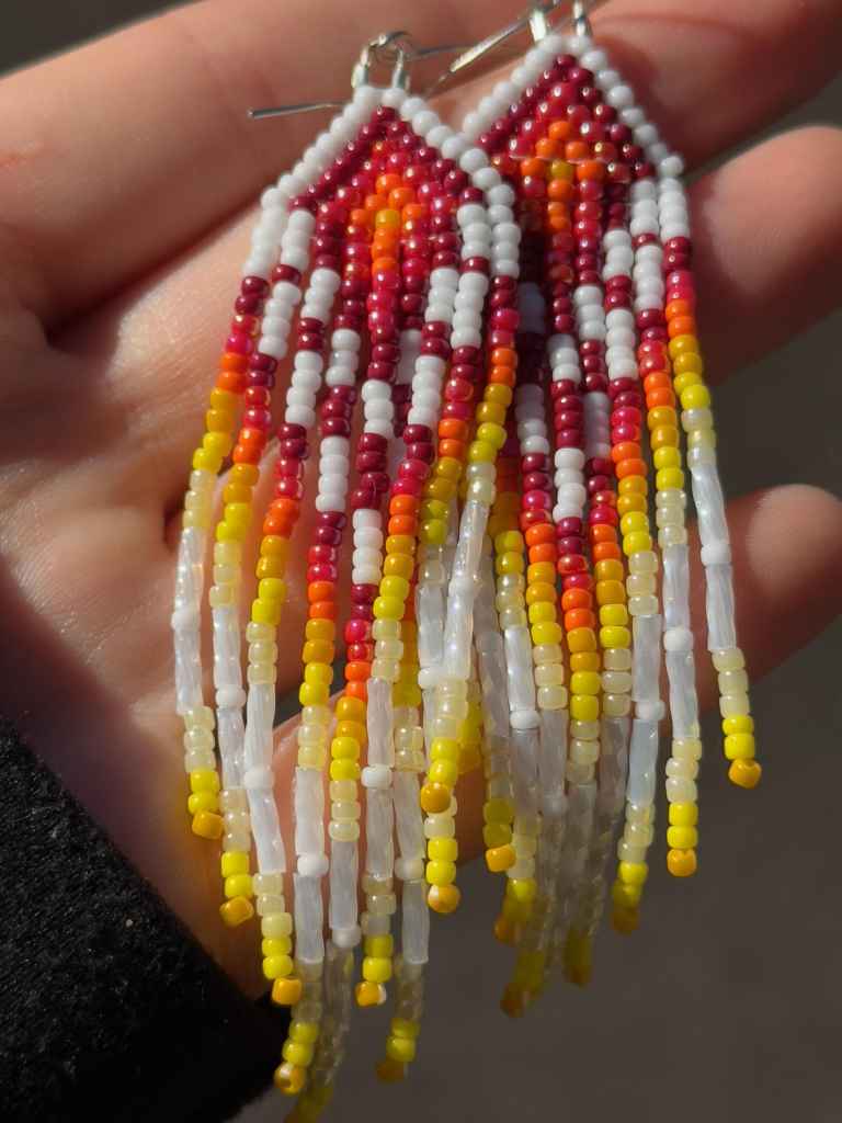 A hand holding a pair of red, orange, yellow, and white beaded fringe earrings. Sunlight hits the luster finish on the red and yellow beads and the white bugle beads.