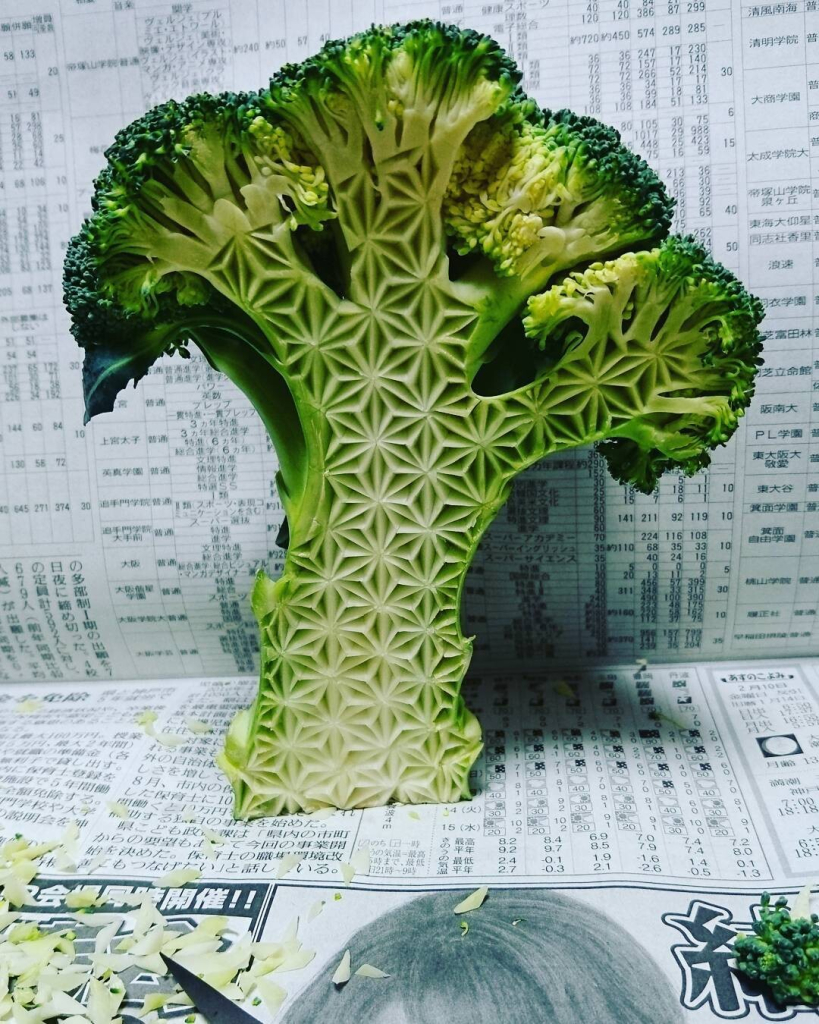 A green piece of broccoli cut in half and carved with intricate, artistic patterns across its side.