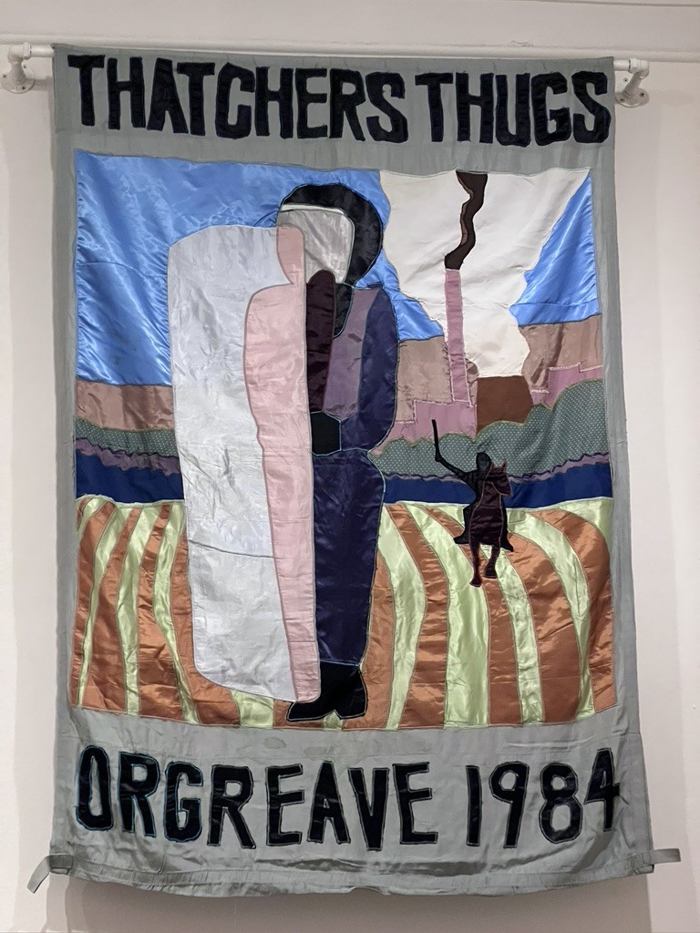 A hanging showing a figure with a shield, presumably a police office, a woman behind and the text ‘Thatchers Thugs, Orgreave 1984’