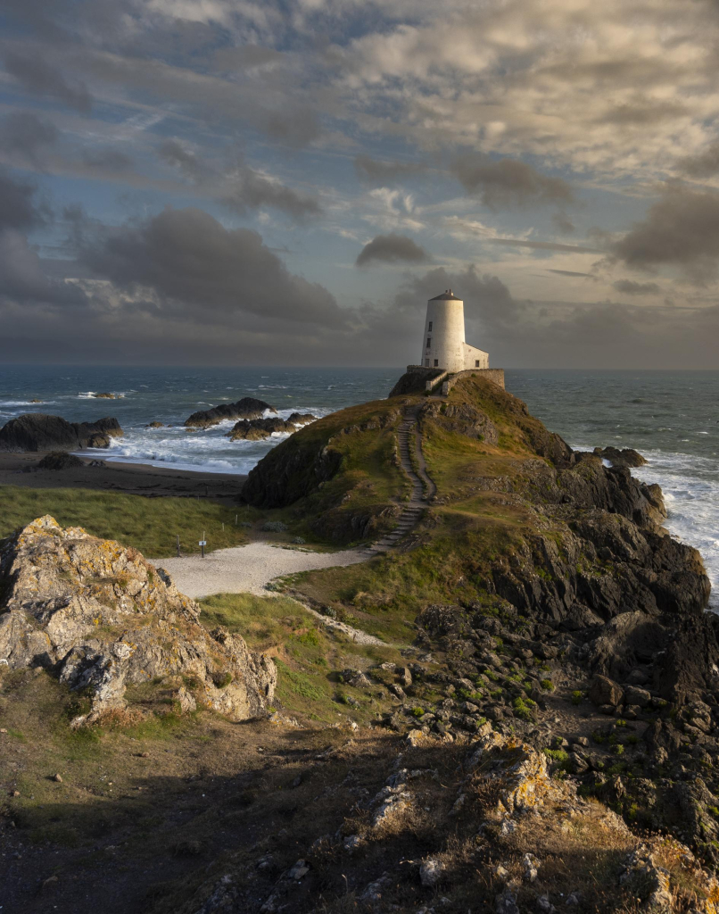 A white painted lighthouse stands on a rocky outcrop next to the sea. Waves crash on the rocks. The sun is setting to the right of the frame casting shadows and giving a golden hue to the scene.