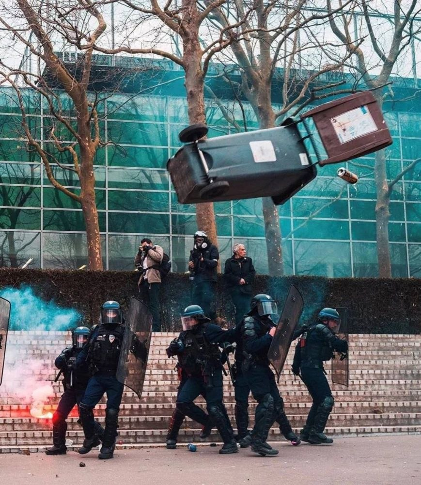 sieben im kreis aufgestellte behelmte und mit schutzschildern versehene Polizist*innen. links im Hintergrund ein kleiner Brennherd. Über den Polizist*innen fliegt eine Mülltönne aus welcher eine Dose fliegt. Im Hintergrund auf einer Mauer (könnte auch eine Treppe sein) ein Mensch mit Kamera, ein/e weitere/r Polizist*in, danbenen ein Mensch welcher zur Seite schaut.