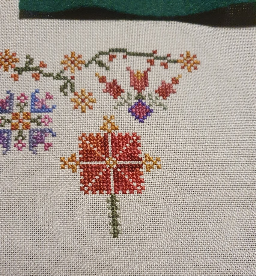 Cross stitched flowers, green vine with yellow flowers along the top, a flower of purples and one of reds underneath the vine. Below that is a square shaped flower in shades of red with golden bits around the outside and a green stem. 