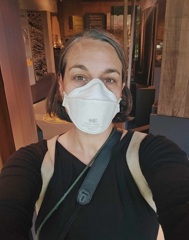 A colour photograph of a woman wearing a white ffp3 mask and looking at the camera. 