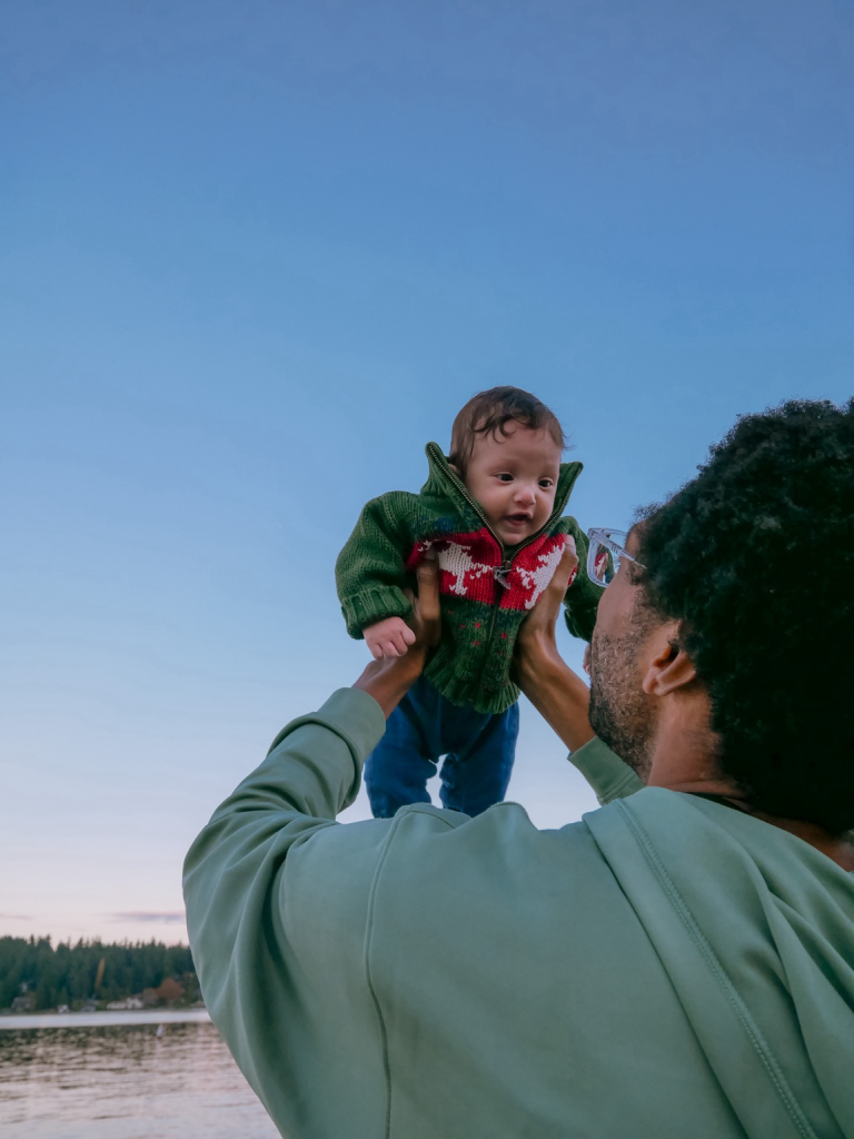 A smiling baby in a green and red sweater being held up to the sky by a Black man facing away from the camera.