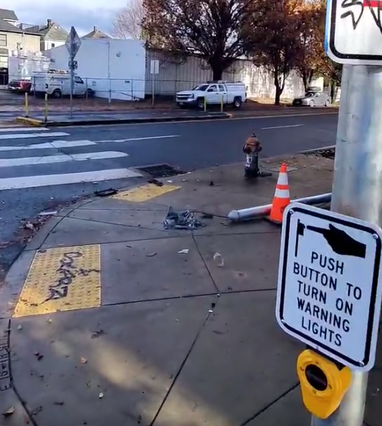 looking right, the sign "push button to turn on warnings lights" is in the foreground and a broken-off warning lights pole laying on the sidewalk behind it.