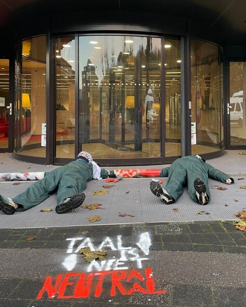 Foto van de ingang van het NOS-gebouw met activisten die met een lock-on voor de draaideur liggen. Op de stoep in krijt: 'TAAL IS NIET NEUTRAAL'