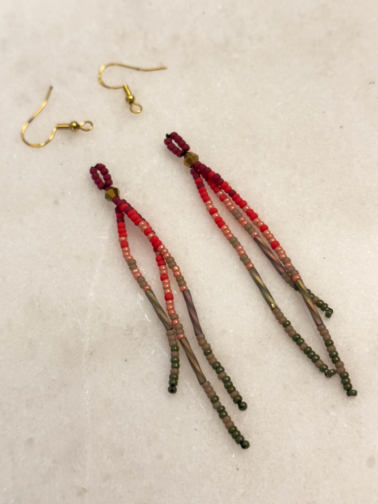 A pair of beaded dangle earrings on a marble background. They fade from dark red, to peach, to dark green.