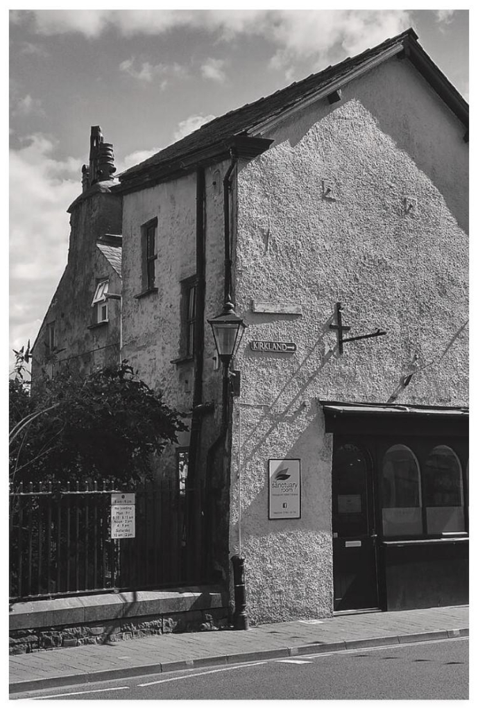 Black and white portrait photograph showing the gable end of a building with an old fashioned Victorian coaching lamp.  The building has a nameplate for Kirkland with an arrow pointing south.