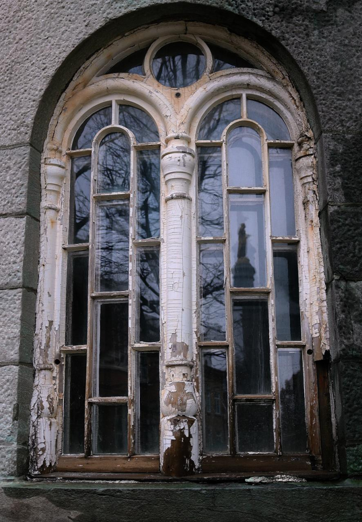 Photograph of an exterior window of St. Jean Baptiste church in Quebec City. The white paint has started to peel, revealing the wooden brown frame. The wall around is of grey stones. A bare tree and blue sky are reflected in the window, as well as the statue of the Saint on a pedestal. Unfortunately this beautiful church was progressively abandoned by its parishioners and is now empty and slowly decaying, to the astonishment of the photographer who lived close by 25 years ago. 