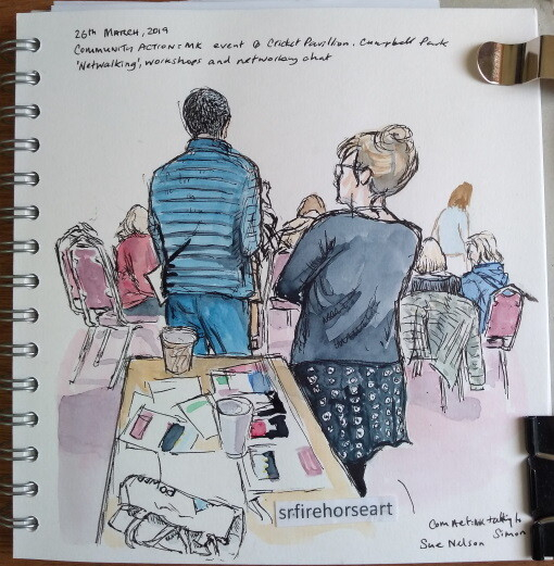 View of a sketchbook page showing a community workshop scene: Two standing figures seen from behind at a community event. In the foreground is a table with leaflets. In the background are  several workshop participants on chairs, also seen from behind. The ink drawing has been painted with watercolours.