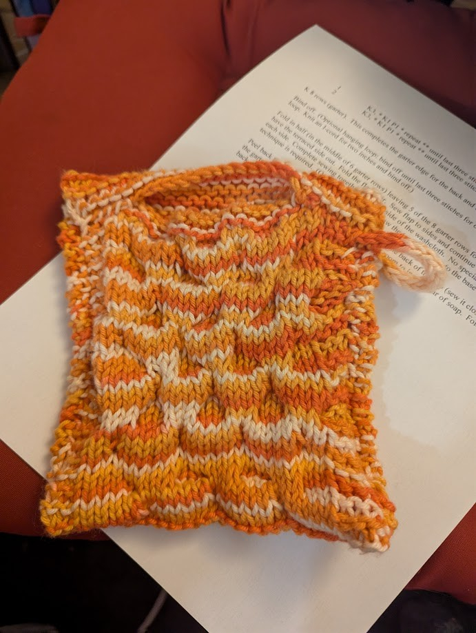 A photo of a finished knitted "soap sock", a knitted "mitt" or pouch meant to hold a bar of soap. The yarn is a bright orange-white mottled colour, and it's got a bubbly knitted texture to it. 