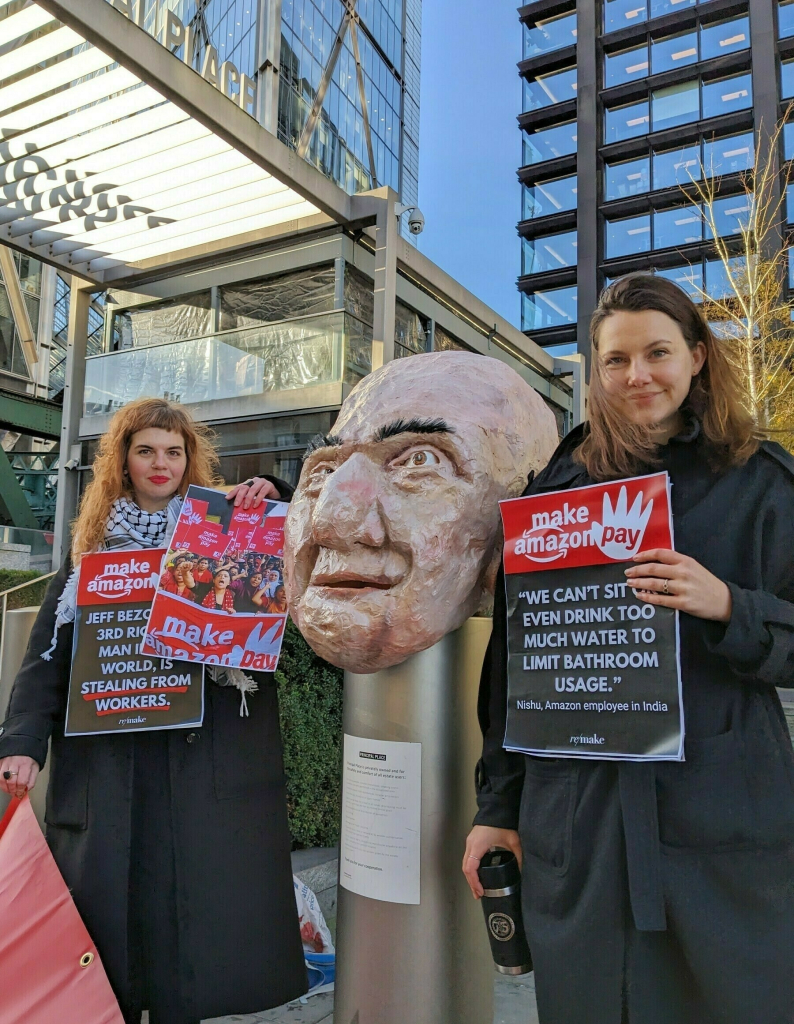 Two women stand beside the paper sculpture of Jeff Bezos's head. They have black coats, long hair and are holding Make Amazon Pay placards