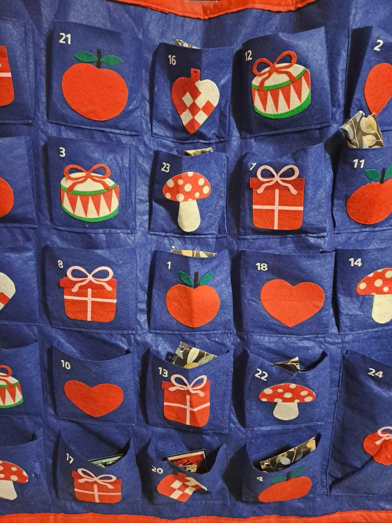 A blue wall hanging with 24 pockets. Each pocket is numbered (at random from 1 to 24), and has simple red, green and white designs on it: apples, hearts, mushrooms, gift boxes etc. The pockets can be filled with whatever one chooses.