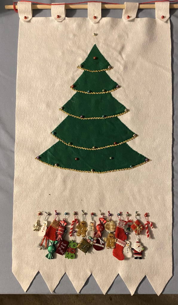 A green felt Christmas tree on a white felt pennant. Small colored beads are sewn into it, creating 24 empty spots on the tree. Below the tree are two rows of similar beads from which hang 24 assorted ornaments that can be put on the tree.