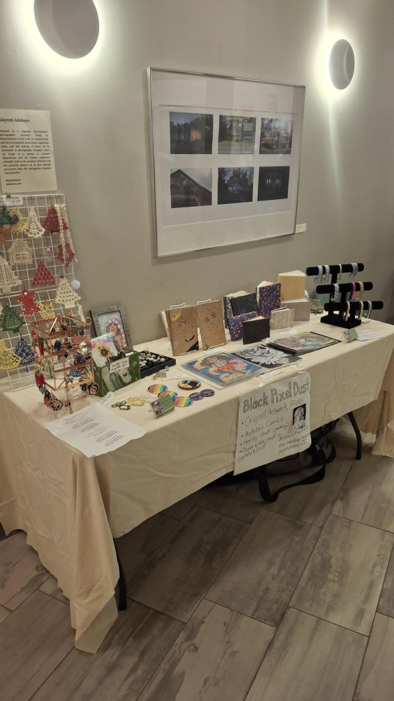 A white table against a white wall. There is a bunch of artwork on the table, handmade earrings, sketchbooks, and beaded jewelry.