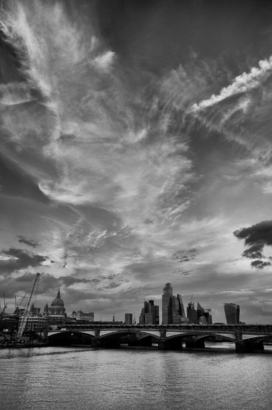 A black and white photograph in portrait orientation of the City of London, taken from the the South Bank in London.
The sky fills the top 80% of the image. It is filled by a variety of wispy clouds in a dramatic fashion.
Below the sky is the skyline of the City. St Paul's Cathedral is prominent on the left of the picture. Several skyscrapers are seen in the bottom centre, stretching to the right, including the (former) NatWest Tower, the "Cheesegrater" and, at the far right, the "Walkie Talkie".
The buildings are interrupted by Blackfriars Bridge crossing the pixture from left to right. Below the bridge is the full width of the River Thames, which looks placid in the evening light.