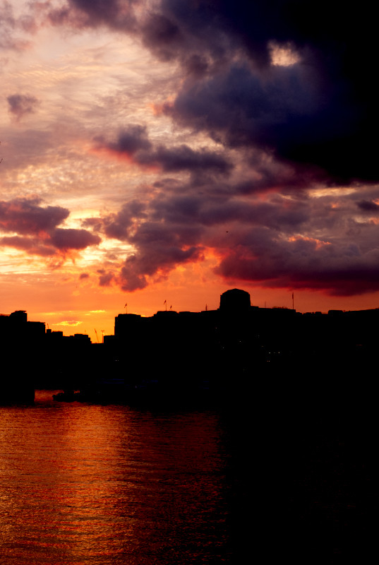 A colour photograph in portrait orientation of sunset over the West End of London, taken from the South Bank.
The sky occupies the top two thirds of the picture. Sky on the right is full of heavy clouds, turned purple by the setting sun. Behind and below them the sky is a fiery red.
The silhouette of buildings on the north shore of the River Thames are seen below the sky, including the tower of the Shell Building.
Below the silhouetted buildings  is the River Thames, which is reflecting the sunset and iss a bright red.