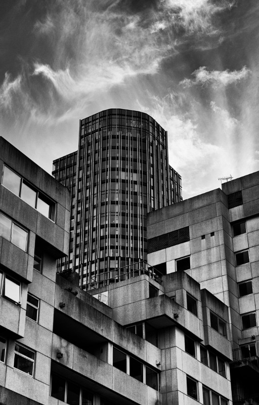 A black and white picture in portrait orientation taken from the Queen's Walk on the South Bank.
It shows some late twentieth century buildings in a brutalist style and a tower block behind. The tower block is a steel-and-glass construction. I believe it's since been demolished - Google Maps shows an empty lot where I believe the tower should be.
Above the buildings is an angry sky full of wispy clouds.