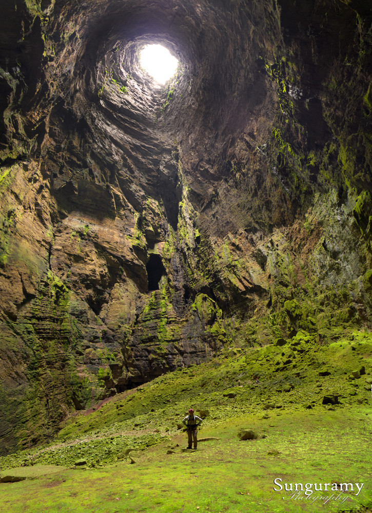 A lone figure stands dwarfed by a massive pit cave, light filtering in from a hole over a thousand feet above their head. The cave is very green with moss and other growth, boulders are the size of houses and cars. The rope we entered on is in this image, but way smaller than a pixel at this distance so it is invisible. In fact, you cannot even see the folk at the base of the rope as the room is that grand. The expanse of the great outdoors, contained underground.