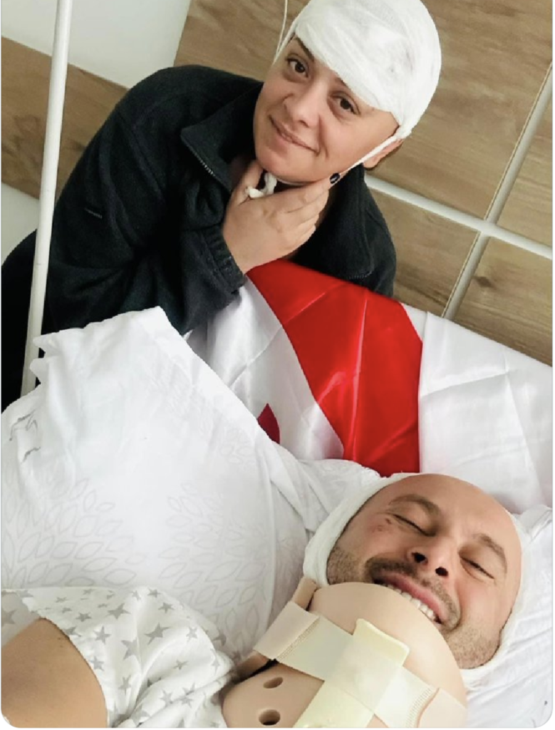 Mariam Gaphrindashvili and Guram Rogava in hospital. Gurum is in a bed with a neck brace and a bandage round his head. Mariam is sat next to him with a bandage on her head. There is a Georgia flag on the bed behind Gurum's head. They are both smiling.