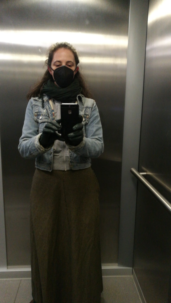 elevator-mirror selfie of a white woman with brown hair in a half-up do. She is wearing a black headstrap respirator, a green scarf, green gloves, a light blue denim jacket a grey hoodie, and an ankle-length skirt with a houndstooth pattern of green, blue, and orange. She is looking down at the phone she is holding out in front of her to take the picture.