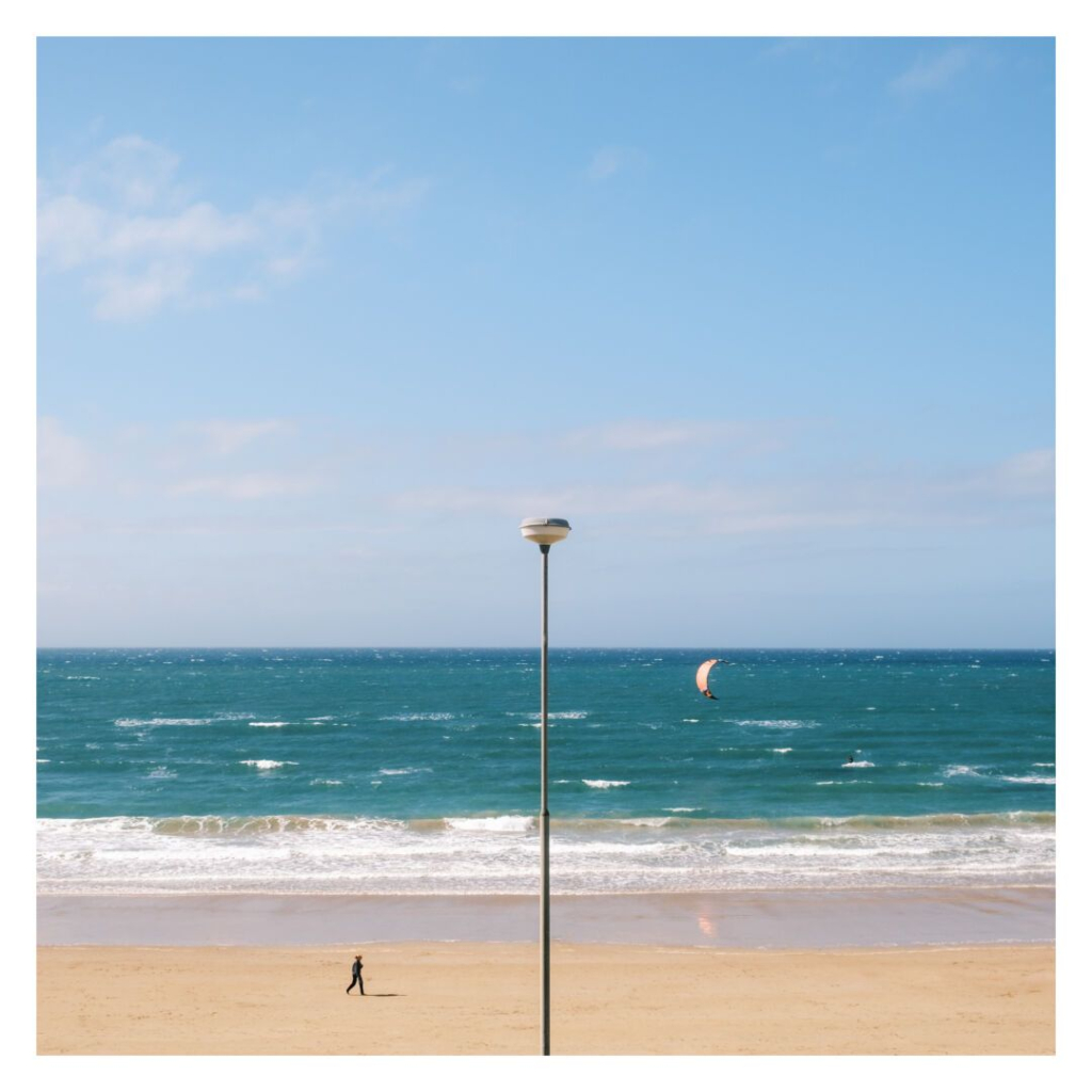 The image is a square color photograph with a white border, depicting a sunny beach scene. The foreground shows a sandy beach with a single person walking on it, wearing a hat, and casting a shadow. In the midground, a lamppost stands tall and centrally placed, dividing the composition. Beyond the beach, the ocean stretches out with small waves visible on its surface. A kitesurfer with an orange kite is visible on the right side of the image, gliding over the water. The sky above is bright blue with scattered clouds, adding to the clear, sunny atmosphere.