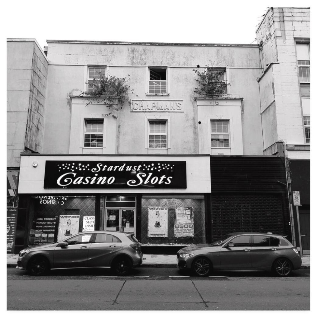 Black and white square photograph showing a weatherworn tatty old high street building with the name Chapman's embossed upon it.  On the ground floor is an amusement arcade signed Stardust Casino Slots.  There are two cars parked in front.