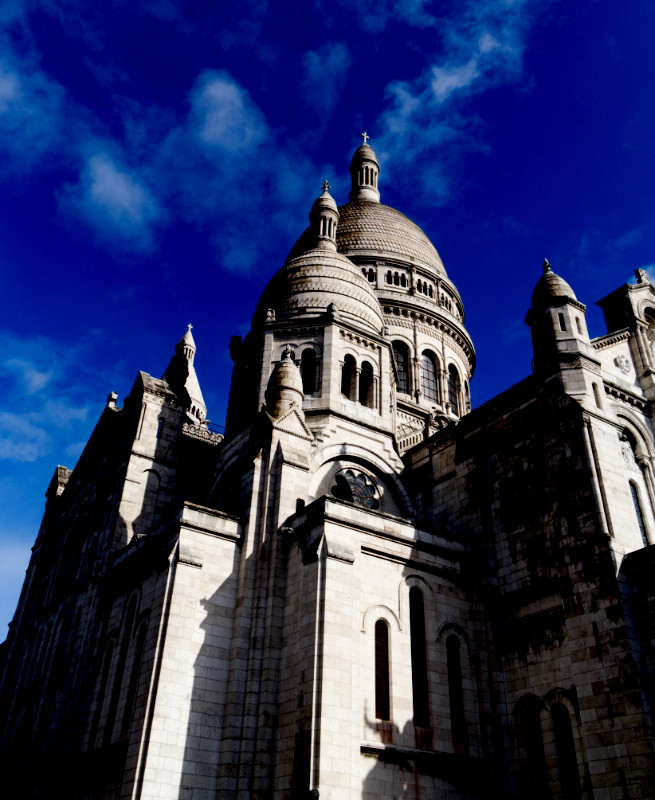 A colour photograph in portrait orientation of the exterior of the  cathedral of Scare Coeur in Montmartre, Paris, on a sunny day. Some bit of the cathedral are strongly illuminated, others are in deep shadow.

The main dome of the cathderal is in the centre of the image, with a lesser dome in front and to the left. There are several spires and towers visible.

Behind the cathedral the sky is an intense deep blue with a few wispy clouds in it.