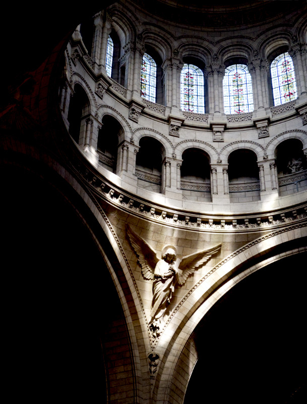 A colour photograph of the interior of the cathedral of Sacre Coeur in Paris, showing the detail of the large main dome.

The picture is centred on a statue of an angel hanging on the triangle of marble formed between two arches supporting the dome.

Above the angel, two further tiers of arches are seen. The top tier of arhces contain windows of stained glass; the arches on the lower tier are filled in with marble.

The arches on each side of the angel are in deep shadow, as is the top of the dome.