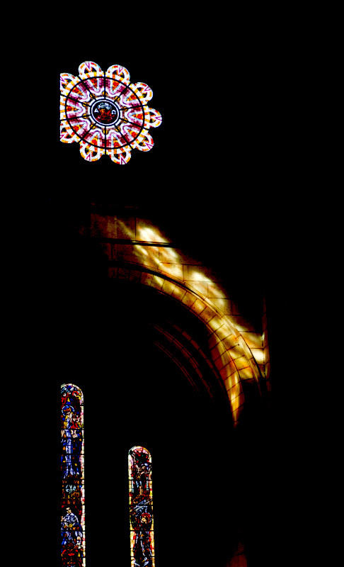 A colour photograph from the interior of the cathedral of Sacre Coeur in Paris.

The interior is in deep, black shadow, with little detail visible. In the top left corner of the image is a brightly lit circular stained glass window. Light from another window illuminates a stone arch below the circular window.

in the bottom left, two further stained glass windows, tall with a rounded top, can be seen. They are full of red and blue light.