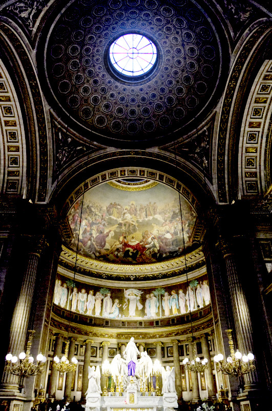 A colour photograph in portrait orientation of the interior of the church of St Madelaine in Paris.

The church is baroque in style, and heavily decorated.

At the top of the picture is the main dome of the church, containing a round window at its summit, and decorated with a repeated pattern of of decreasing circles.

The dome is supported on arches, one to the rear over the apse (in the centre of the picture) and one on each side. Beneath the main arch, the apse is decorated by a painting on the curved ceiling of another dome. The painting shows a detailed biblical crowd scene, possibly the sermon on the mount.

Beneath the painted dome is a band relief of many people, possibly saints.

In the apse is an altar, decorated with a detail baroque scuplture of Christ.