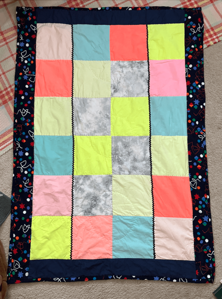 A simple quilt made from twenty-four squares in neon colors, arranged in a 6x4 rectangle; a dark print fabric forms the border. There are two rows of black rickrack running the long way through the body of the quilt.